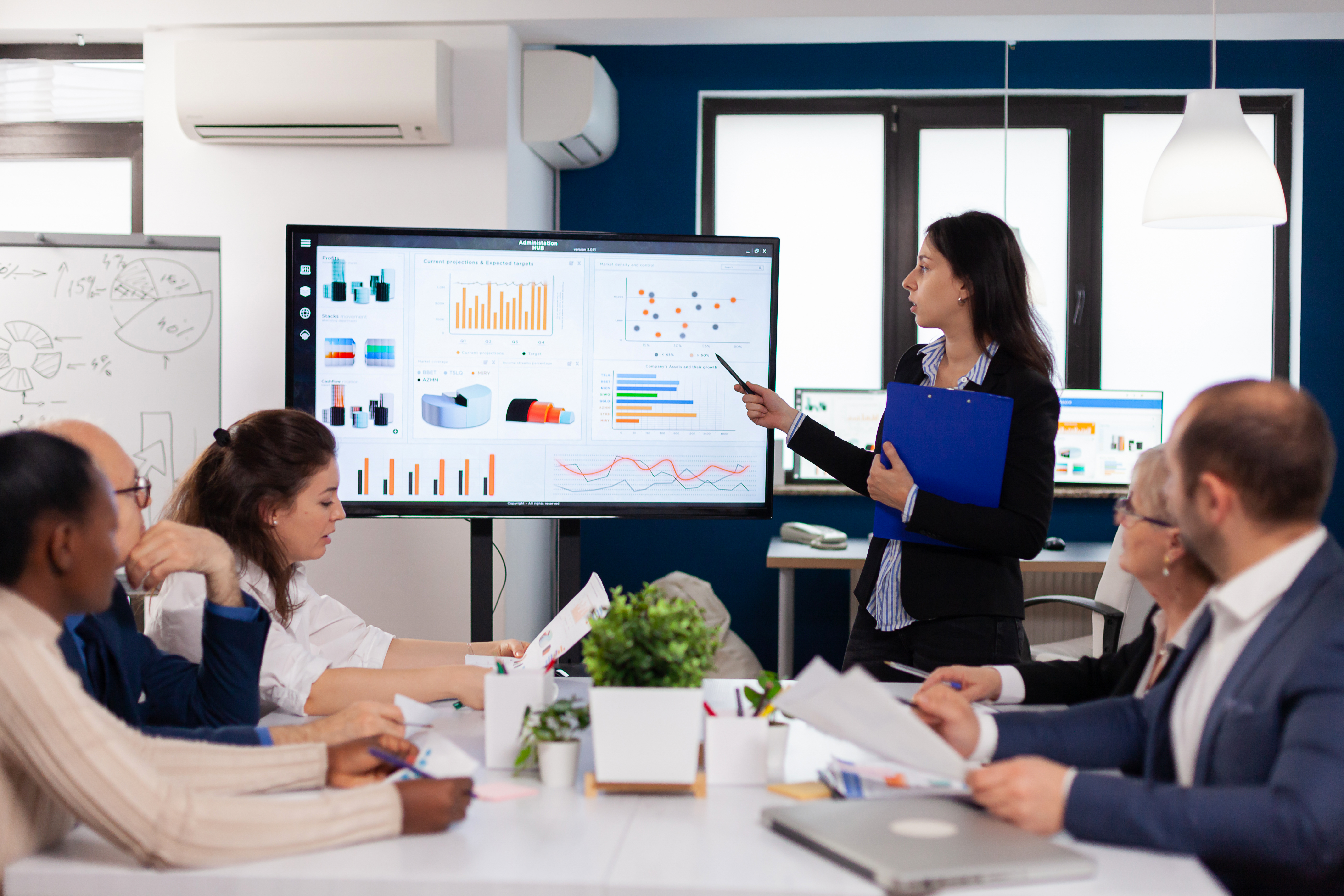 Young team leader in big corporation briefing coworkers pointing at graph meeting. Corporate staff discussing new business application with colleagues looking at screen